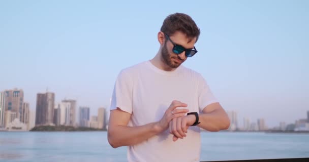 Un joven de pie en el paseo marítimo en el verano utiliza la pantalla de reloj inteligente — Vídeo de stock
