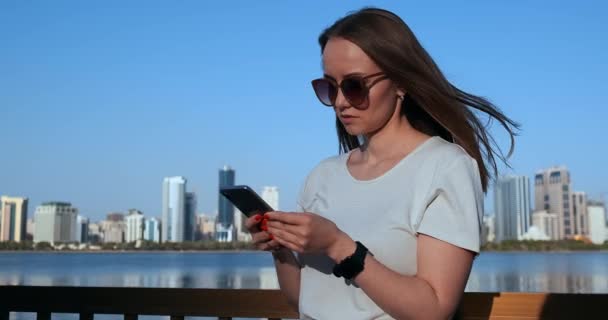 Девушка с длинными волосами набирает сообщение на смартфоне на набережной Дубая . — стоковое видео