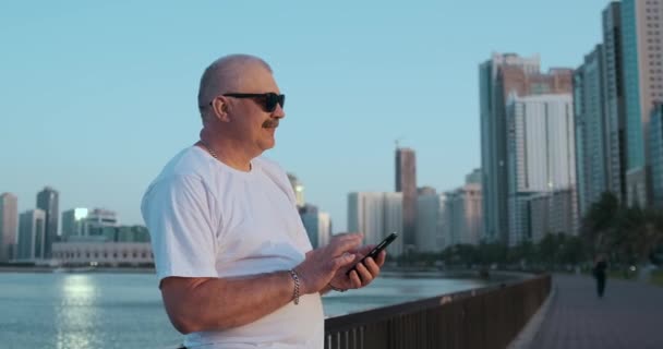 Hombre mayor barbudo parado al aire libre usando un teléfono inteligente. Hombre retirado leyendo noticias, charlando con sus hijos a través del teléfono móvil. Estudiar la aplicación mapa de la ciudad. Perdido en la ciudad buscando la dirección correcta — Vídeos de Stock