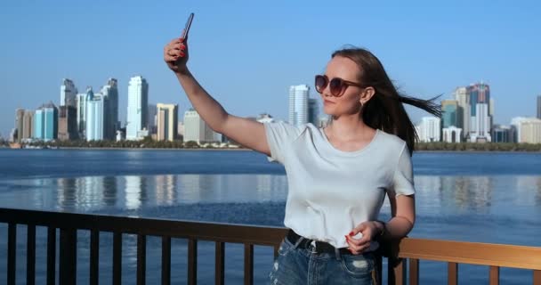Caucasian woman is taking selfies by mobile phone, outdoors in Dubai Marina in sunny day — Stock Video