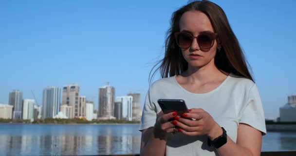 Bella ragazza con i capelli lunghi in occhiali da sole utilizzando app smartphone al molo del fiume tramonto vicino 4K . — Video Stock