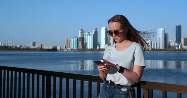 Hermosa chica con el pelo largo en gafas de sol usando aplicación de teléfono inteligente en el muelle del río puesta de sol cerca de 4K . — Vídeos de Stock