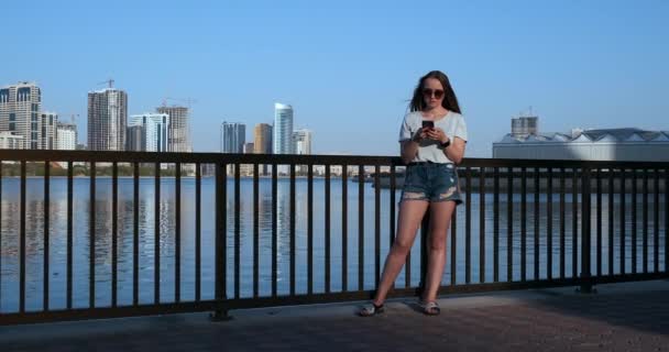 Schöne Mädchen mit langen Haaren in Sonnenbrille mit Smartphone-App am Sonnenuntergang River Quay in der Nähe 4k. — Stockvideo