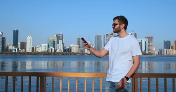 Ένας άντρας το καλοκαίρι στέκεται στην προκυμαία φωτογραφημένος σε ένα smartphone τοπίο της πόλης — Αρχείο Βίντεο