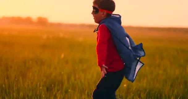 Een jongen in een superheld kostuum bij zonsondergang loopt over het veld lachen en glimlachen — Stockvideo