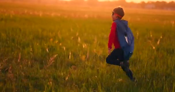 Un niño con un traje y una máscara de superhéroe corriendo por el campo al atardecer en la hierba — Vídeos de Stock