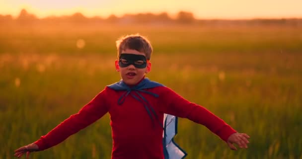 Un niño con un traje y una máscara de superhéroe corriendo por el campo al atardecer en la hierba — Vídeos de Stock