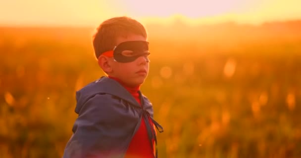 Un niño disfrazado de superhéroe con una capa roja atraviesa el césped verde con el telón de fondo de una puesta de sol hacia la cámara — Vídeos de Stock