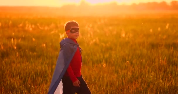 Un bambino in costume da supereroe con un mantello rosso corre attraverso il prato verde sullo sfondo di un tramonto verso la telecamera — Video Stock