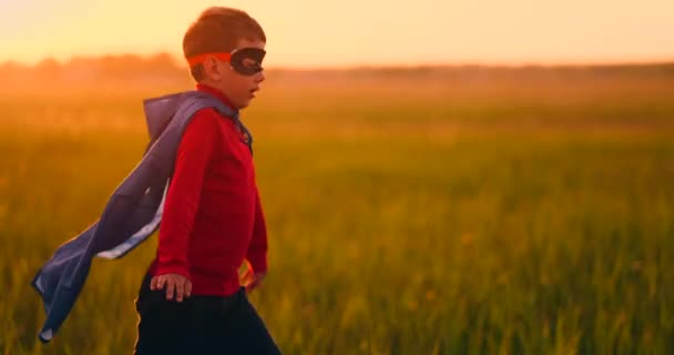 Un niño con un disfraz de superhéroe al atardecer corre por el campo riendo y sonriendo — Vídeos de Stock