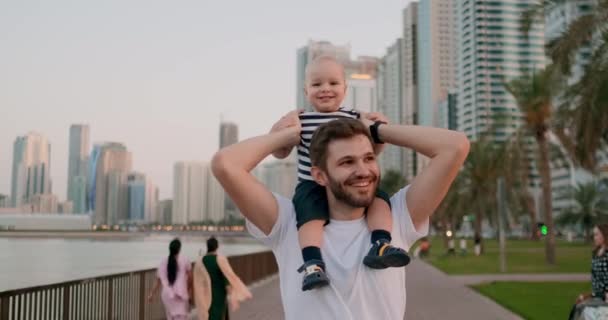 Улыбающийся молодой отец гуляет с ребенком, сидящим на шее летом, город рядом с водой и пальмами — стоковое видео