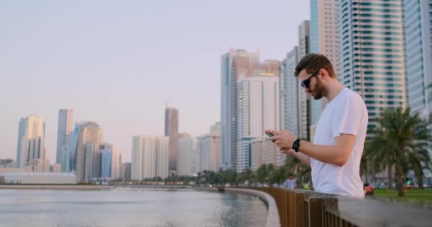 Man tourist standing on the waterfront writing text messages on your smartphone — Stock Video