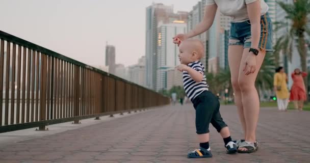 Smiling happy laughing baby takes first steps in summer on the waterfront with mom pointing her finger — Stock Video
