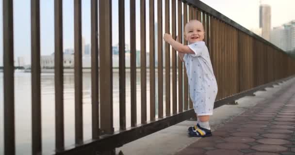 Bailando alegre chico europeo cerca de la valla mantiene ha barandilla y baile de pie y mirando a la vista del paseo marítimo en el verano en los Emiratos Árabes Unidos — Vídeo de stock