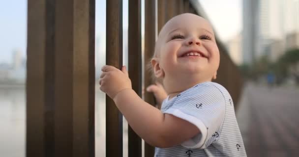 Der Junge lacht und blickt in die Kamera, die im Sommer am Ufer steht und das Geländer hält — Stockvideo