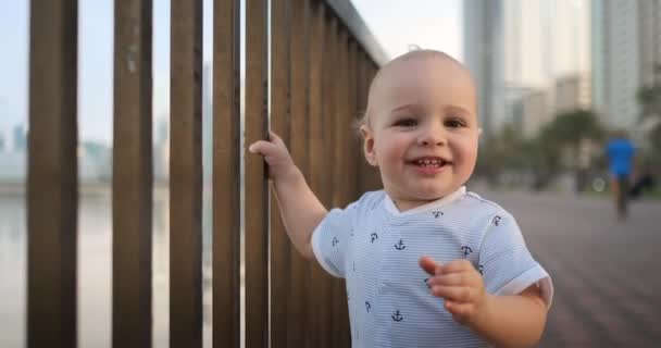 Смеющийся мальчик в возрасте 1 года танцует держа перила и учится ходить делая первые шаги стоя в белом комбинезоне на набережной — стоковое видео