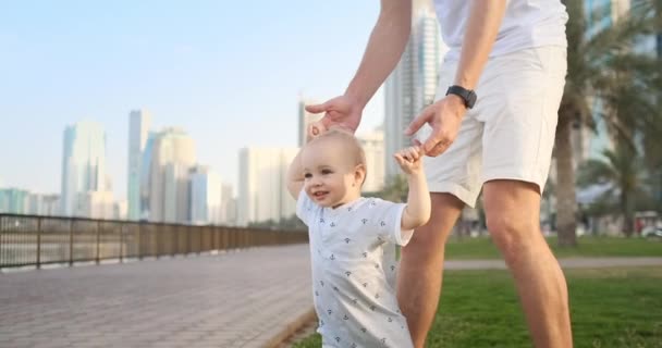 Joven padre con un hijo y los primeros pasos. Padre joven con un niño en el aprendizaje al aire libre para un primer paso cerca de la ciudad urbana — Vídeo de stock