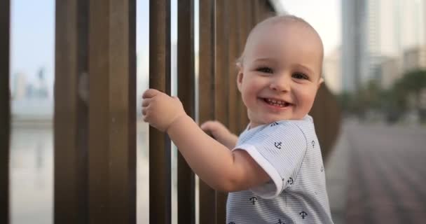 Chlapec se směje a dívá se na kameru stojící na nábřeží v létě, kde se drží zábradlí — Stock video