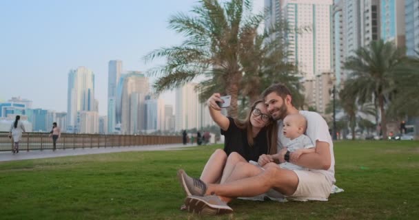 Parkta çim üzerinde birlikte oturan ve bir selfie alarak iki çocuk ile mutlu bir aile. Akıllı telefon ile — Stok video