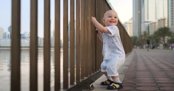 Śmiech chłopiec w wieku 1 roku taniec trzyma poręcze i uczenia się chodzić podejmowania pierwszych kroków stojących w białym kombinezon na nabrzeżu — Wideo stockowe