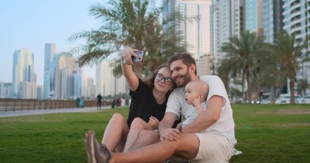 Familia feliz con dos niños sentados juntos en el césped en el parque y tomando una selfie. Con smartphone — Vídeo de stock