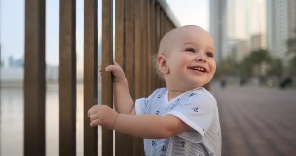 Хлопчик, який сміється у віці 1 року, танцює, тримає перила і вчиться ходити, роблячи перші кроки, що стоять у білому комбінезоні на набережній — стокове відео