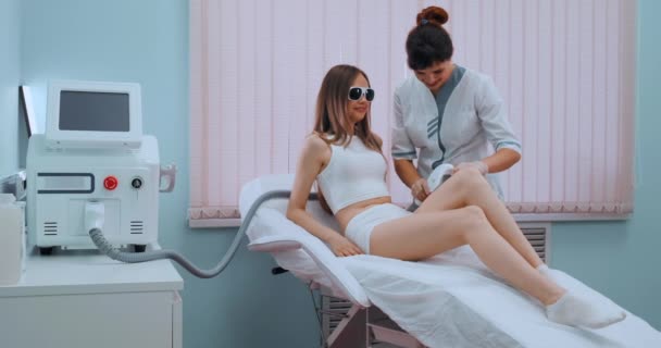 Épilation laser sur les jambes des femmes. La fille est allongée sur le canapé dans les lunettes médicales dans la salle de traitement. Les mains du cosmétologue font l'épilation des jambes . — Video