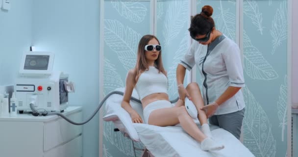 A mulher veio ao procedimento da remoção do cabelo do laser. O médico trata sua coxa com um dispositivo — Vídeo de Stock