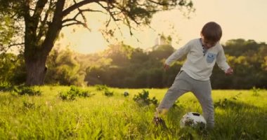 Yavaş çekimde, çocuk gün batımında bir çayırda bir futbol topu ile komik oynar.