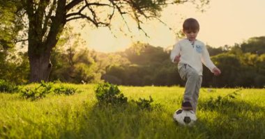 Yavaş çekimde, çocuk gün batımında bir çayırda bir futbol topu ile komik oynar.