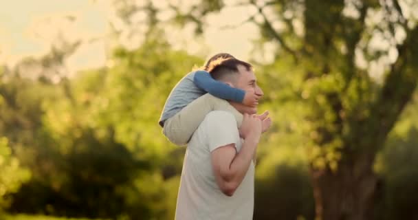 Un niño sentado en el cuello de su padre mientras camina en el campo de verano al atardecer — Vídeo de stock