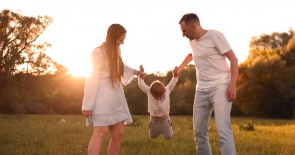 Maminka a táta potřásli dítěti, jak si hrají na rukách, chlapec je šťastný, když si hraje s rodiči na ulici v létě při západu slunce. — Stock video