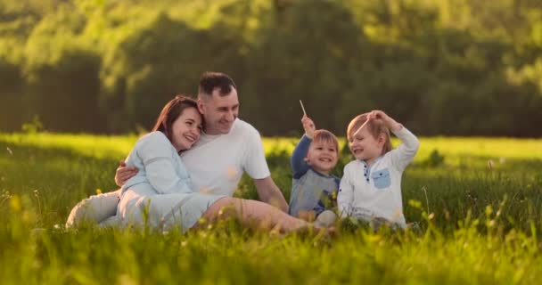 Papá mamá y dos hijos en el verano al atardecer sentados en un prado en la hierba riendo y abrazándose — Vídeo de stock