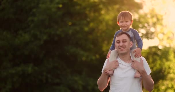 Ο στοργικός πατέρας χαμογελά περπατώντας με το παιδί που κάθεται στο λαιμό στο ηλιοβασίλεμα σε ένα λιβάδι το καλοκαίρι σε αργή κίνηση — Αρχείο Βίντεο
