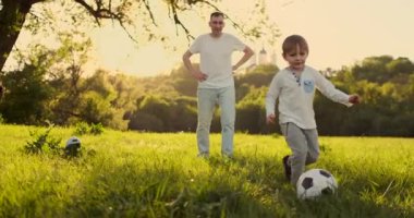 Baba gün batımında oğluyla futbol oynarken kapıda duruyor. Baba çocuğu sahada eğitir. Çocuk topa vurur.