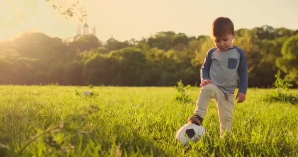 Мальчик стоит на траве с футбольным мячом на закате — стоковое видео