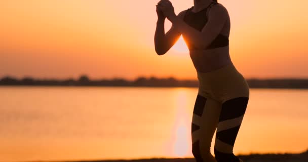 Boční pohled na mladou ženu, která dělá squaty venku. Pohled na mladou ženu s posilováním, stojící u jezera na písku při západu slunce, pomalý pohyb. — Stock video