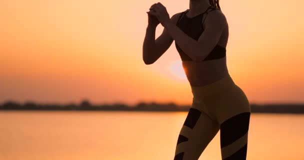 Seitenansicht einer jungen Frau, die im Freien Kniebeugen macht. Seitenansicht einer jungen Fitness-Frau beim Kniebeugen im Stehen am See im Sand bei Sonnenuntergang in Zeitlupe. — Stockvideo
