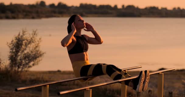 Magmusklerna övningar. En ung skönhet atletisk i solnedgången mot sjön i sommar kvinna i sportkläder gör övningar för ABS i slow motion på baren. — Stockvideo