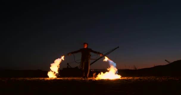 En man i en regnrock med två eldkastare släpper ut en eldig flamma stående vid solnedgången på sanden. — Stockvideo