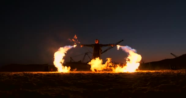 Człowiek w płaszczu przeciwdeszczowym z dwoma miotaczami ognia pozwala na ognisty płomień stojący o zachodzie słońca na piasku. — Wideo stockowe