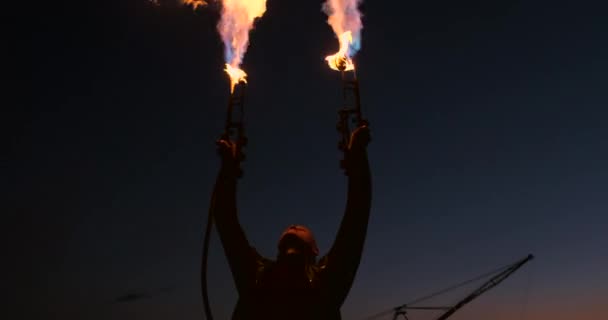 Ein Mann im Regenmantel mit zwei Flammenwerfern lässt eine feurige Flamme im Sonnenuntergang auf dem Sand stehen. — Stockvideo