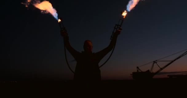 2つの火炎放射器を持つレインコートの男は、砂の上に日没時に立って燃える炎を出します. — ストック動画