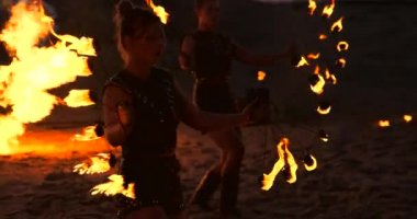 Yağmurluklu, iki alev makinesi olan bir adam gün batımında kumda duran ateşli bir alev salar..