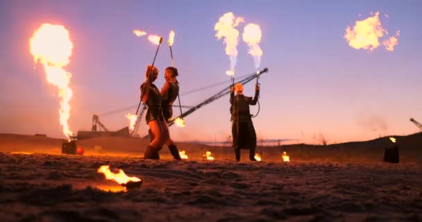 Um grupo de homens e mulheres show de fogo à noite na areia contra o fundo do fogo e guindastes torre — Vídeo de Stock