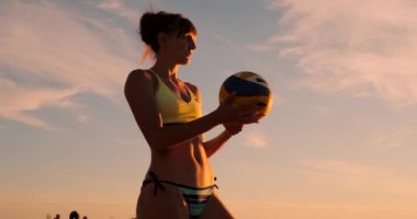 Gün batımında bir top ile bikini güzel bir kadın kum üzerinde bir voleybol maçında plajda atlama hizmet yapmaya hazırlanıyor.