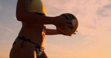 Gün batımında bir top ile bikini güzel bir kadın kum üzerinde bir voleybol maçında plajda atlama hizmet yapmaya hazırlanıyor.