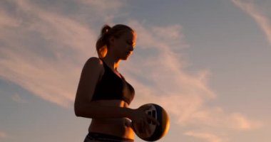 Gün batımında bir top ile bikini güzel bir kadın kum üzerinde bir voleybol maçında plajda atlama hizmet yapmaya hazırlanıyor.