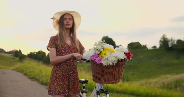 Middelste plan meisje in jurk gaat met fiets en bloemen in het veld. — Stockvideo