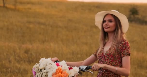 Porter girl marche sur la caméra dans une robe avec des fleurs dans un panier et un vélo dans le champ . — Video